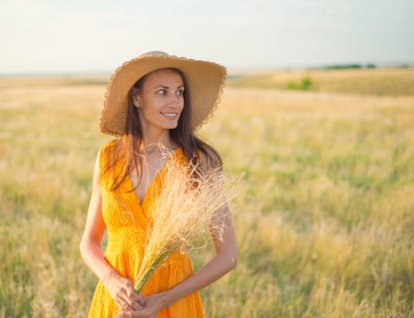 Young woman in an orange dress and a straw hat standing on a field in the rays of the setting sun.