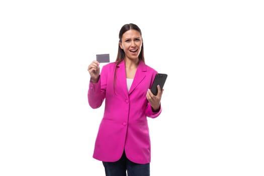 young pretty woman with black straight hair is dressed in a trendy pink jacket holding a credit card and a smartphone.