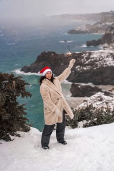 Outdoor winter portrait of happy smiling woman, light faux fur coat holding heart sparkler, posing against sea and snow background.