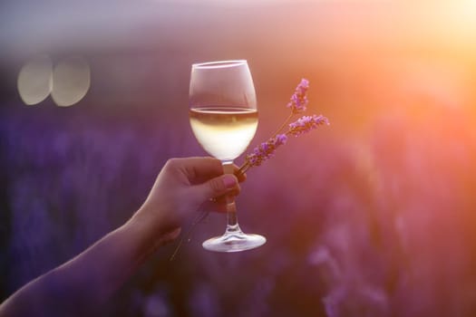 Glass white wine lavender field. Woman hand holds a glass with lavander and wine in the Lavender field at sunset Violet flowers on the background.. Conscious consumption. Wellness and natural concept.