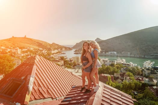 women standing on rooftop, enjoys town view and sea mountains. Peaceful rooftop relaxation. Below her, there is a town with several boats visible in the water. Rooftop vantage point