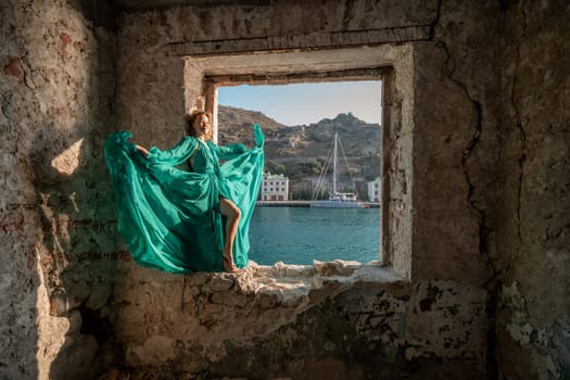 Rear view of a happy blonde woman in a long mint dress posing against the backdrop of the sea in an old building with columns. Girl in nature against the blue sky