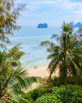 View between palm tree leaves at a couple on the beach, Drone aerial view at Koh Ngai island with palm trees and soft white sand, and a turqouse colored ocean in Koh Ngai Trang Thailand