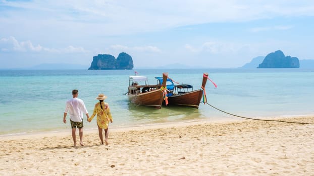 a couple of men and woman walking on the beach with longtail boats in the ocean of Koh Ngai island soft white sand, and a turqouse colored ocean in Koh Ngai Trang Thailand