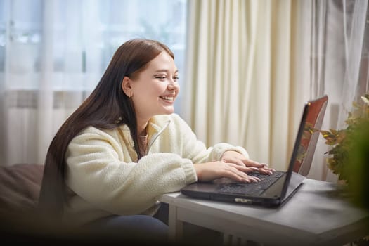 Fashionable young overweight plump female freelancer with portable computer, typing, working remotely from home. Brunette plus size girl using laptop