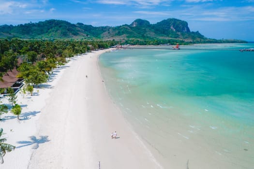 Drone view at a couple walking on the white sandy tropical beach of Koh Muk with palm trees soft white sand, and a turqouse colored ocean in Koh Mook Trang Thailand