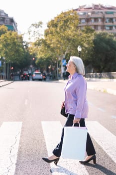 senior woman crossing a city street at a pedestrian crossing with shopping bags in her hand, concept of elderly people leisure and active lifestyle
