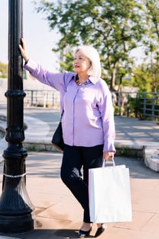 senior woman waiting standing leaning on a lamppost with shopping bags in her hand, concept of elderly people leisure and active lifestyle