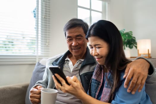 Retired elderly couple sits on couch drink tea and using mobile together and relax in their home. Senior Activity Concept.