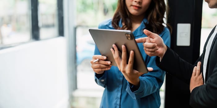 Asian woman and business man working using digital tablet tech discussing financial market data standing at corporate office.