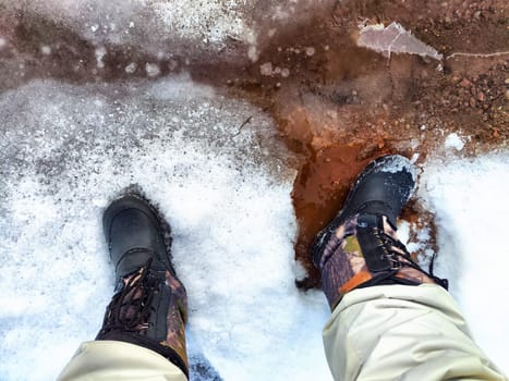 Feet of Hunter or fisherman in big warm boots on winter day on snow. Top view. Fisherman on the ice of a river, lake, reservoir on spring day with melting ice. Dangerous fishing in autumn