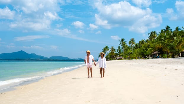 a young couple of caucasian men and a Thai Asian woman walking at the beach o Koh Muk a tropical island, with palm trees soft white sand, and a turqouse colored ocean in Koh Mook Trang Thailand