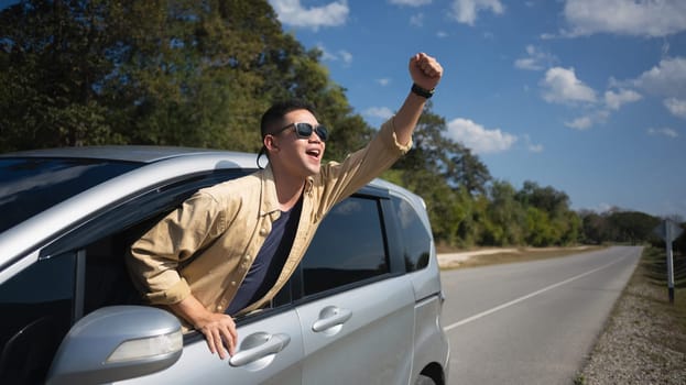 Excited man leaning out of car window enjoying nature on country road. Travel and active lifestyle concept.