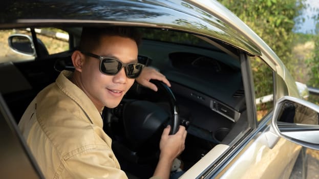 Happy young Asian man in sunglasses driving car in countryside road while traveling alone.