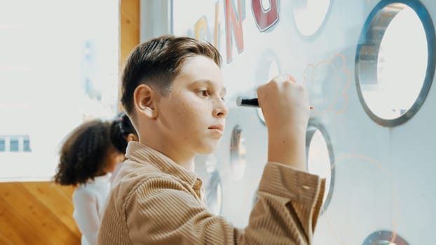 Closeup of caucasian creative student draw beautiful picture at glass board in art lesson. Smart boy using marker drawing while multicultural friend drafting together. Creativity education. Erudition.