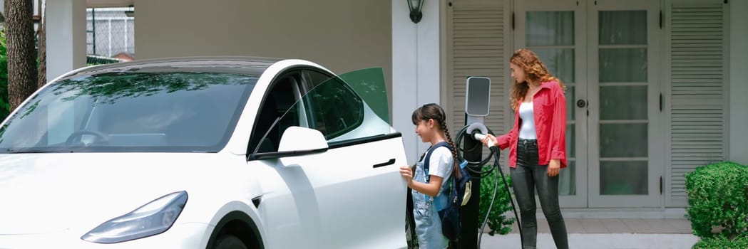 Happy little young girl learn about eco-friendly and energy sustainability as she help her mother recharge electric vehicle from home EV charging station. EV car and modern family. Panorama Synchronos