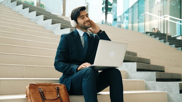 Professional business man sitting at stairs while working on laptop. Skilled project manager listening music from headphone and checking email and discussion about marketing plan. Outdoor. Exultant.