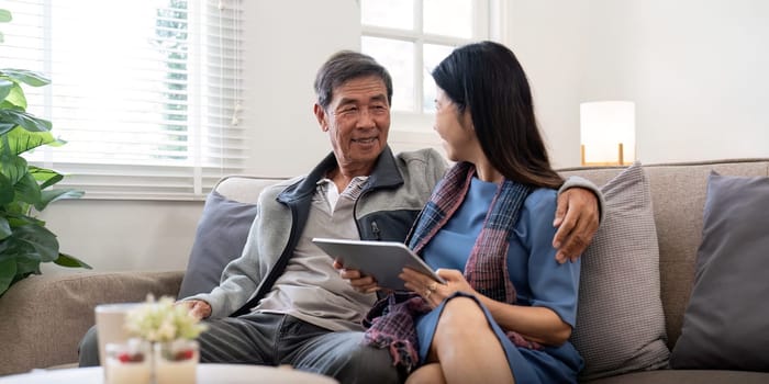 Happy elderly asian couple using tablet sit on sofa doing ecommerce shopping online on website and buying insurance browsing at home.