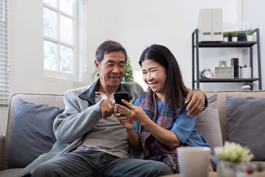 Happy elderly asian couple using smartphone sit on sofa doing ecommerce shopping online on website at home.