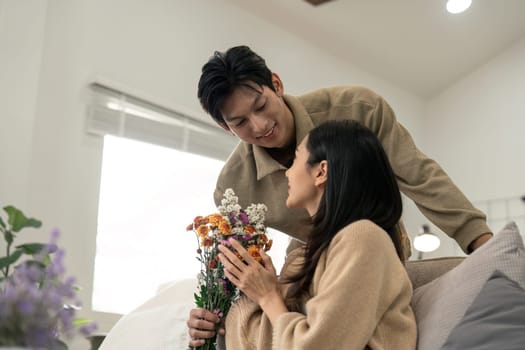 Romantic young asian couple embracing with holding flowers and smiling in living room at home. fall in love. Valentine concept.