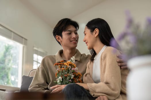 Romantic young asian couple embracing with holding flowers and smiling in living room at home. fall in love. Valentine concept.