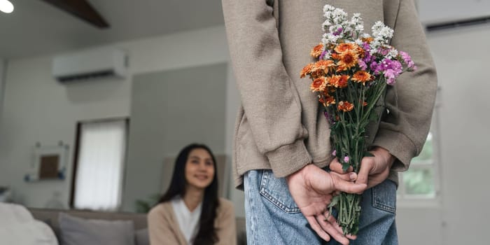 Romantic young asian couple embracing with holding flowers and smiling in living room at home. fall in love. Valentine concept.