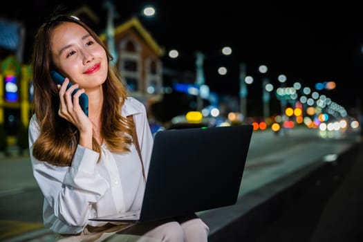 A woman is hard at work on a busy city street corner at night, multitasking with her laptop and cell phone to maximize her productivity and stay connected in the digital age.