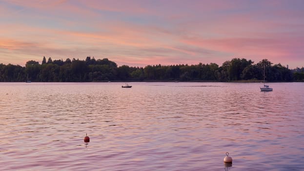 Bodensee Lake Sunrise Panorama. Morning Sunlight Over Tranquil Waters. Witness the mesmerizing dawn over Germany's Bodensee Lake, captured from a boat dock. Embrace the tranquil beauty of the early morning as the sun rises, casting a soft glow on the landscape. The peaceful scene features boats, yachts, and a charming water shack set against a backdrop of a captivating sky. Clouds delicately reflect on the calm water, creating a serene atmosphere. Immerse yourself in the serene beauty of a lakeside sunrise. Explore the harmony of nature, technology, and production as the day unfolds by the lake.