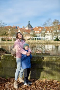 Riverside Family Harmony: Mother, 30 Years Old, and Son - Beautiful 8-Year-Old Boy, Standing by Neckar River and Historic Half-Timbered Town, Bietigheim-Bissingen, Germany, Autumn. Immerse yourself in the picturesque harmony of familial bonds by the riverside with this captivating image. A mother, 30 years old, and her son - a beautiful 8-year-old boy, standing by Neckar River and the historic half-timbered town of Bietigheim-Bissingen, Germany. The autumnal colors add warmth to this timeless family moment.