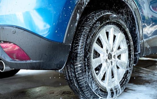 Blue car wash with white soap foam and professional auto care service. Car cleaning service concept. Vehicle cleaning service. Foam wash car detailing. Tire of car wheel is covered with white foam.