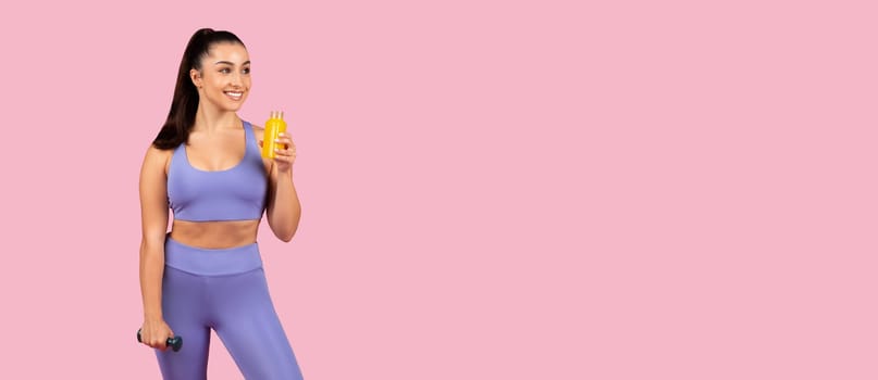 Radiant fitness model in purple sportswear holding yellow juice bottle and green dumbbell, promoting healthy lifestyle on pink backdrop, panorama with free space