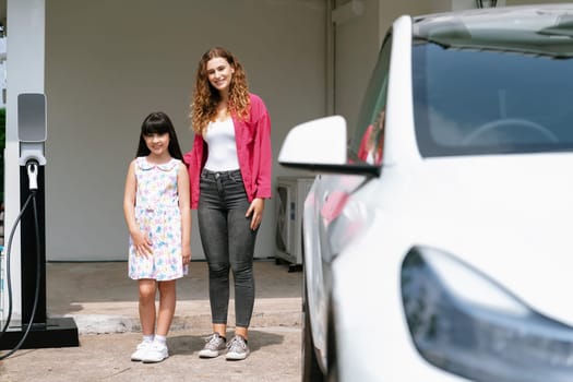 Happy little young girl learn about eco-friendly and energy sustainability as she help her mother recharge electric vehicle from home EV charging station. EV car and modern family concept. Synchronos