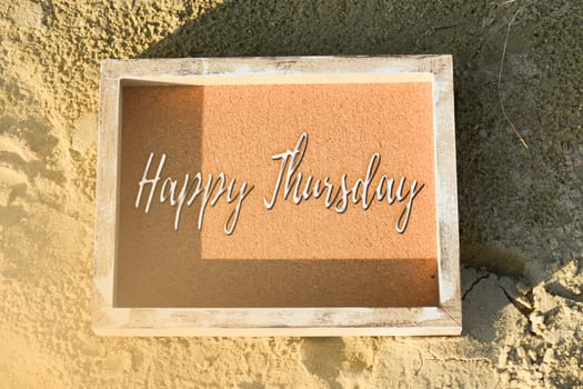 Happy Thursday text on wooden frame laying on sandy beach.