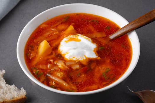 Close up borscht with sour cream in a bowl with bread on a gray table.