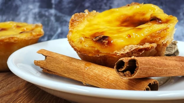 One Pastel de nata or Portuguese egg tart and cinnamon sticks on a white plate. Pastel de Belm is a small pie with a crispy puff pastry crust and a custard cream filling