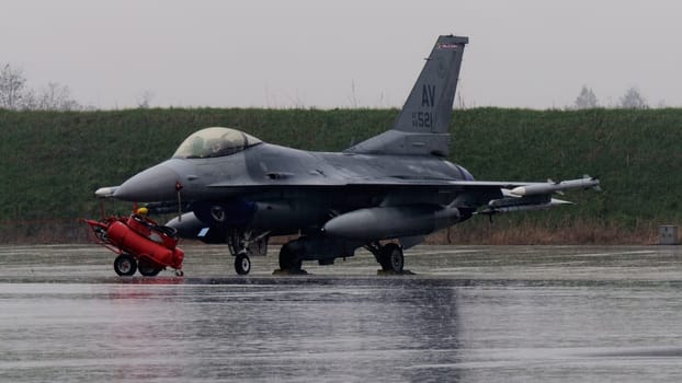 United States F-16 defense aircraft, armed waiting takeoff under the rain, symbolizes NATO unwavering commitment to airspace security, regardless of weather conditions.