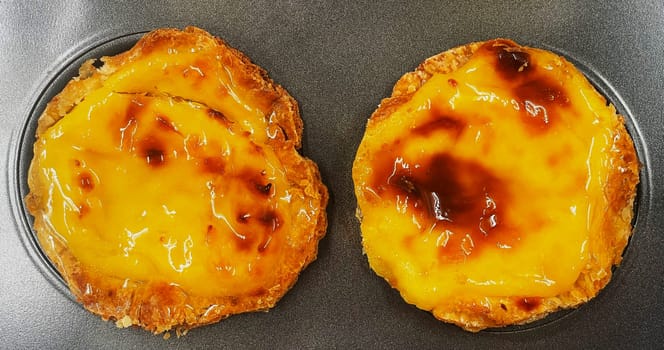 Two freshly baked Pastel de nata or Portuguese egg tart desserts in a baking dish. Pastel de Belm is a small pie with a crispy puff pastry crust and a custard cream filling. A small dessert, a cupcake