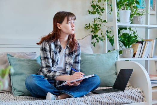 College student girl studying at home, sitting on the couch with laptop, making notes in study book. Education, training, technology, youth concept