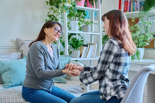 Talking mother and teenage daughter sitting together on couch at home, smiling holding hands. Family, communication, motherhood, friendship, relationship between parent and daughter 18-20 years old