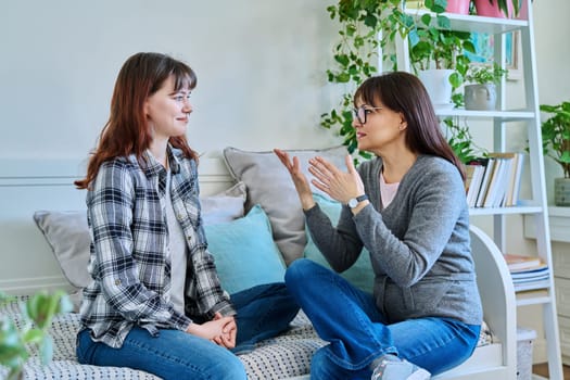 Talking happy smiling mother and teenage daughter sitting together on couch at home. Family, communication, motherhood, friendship, relationship between parent and daughter 18-20 years old