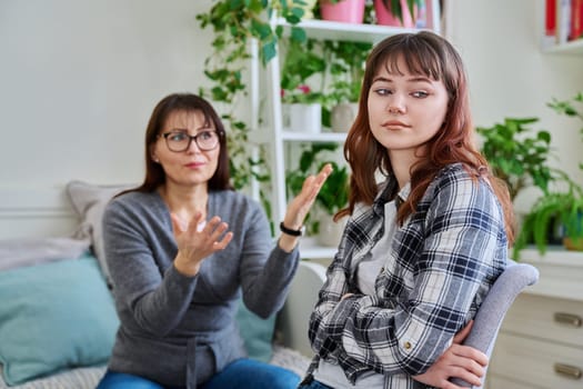 Conflict between mother and teen daughter, offended teenage girl sitting with back to upset arguing mother, on couch at home. Family, difficulties, problems, differences between two generations