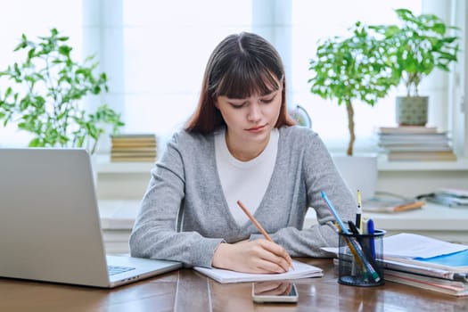 College student girl sitting at desk using laptop computer, making notes in study notebook, at home. Teenager female watching webinar, preparing for exam tests, studying remotely.