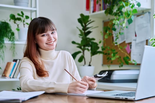 Young female college student studying at home at desk using computer laptop, writing in notebook, smiling looking at camera. E-learning, education, technology, knowledge, youth concept