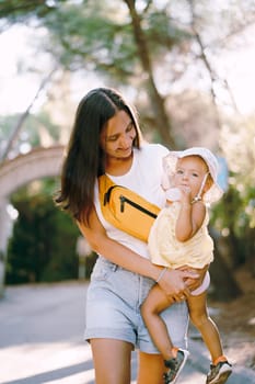 Mom with a little girl in her arms, drinking water from a bottle, walks along the road in the park. High quality photo