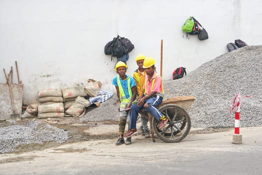 Antananarivo, Madagascar - April 24, 2019: Group of unknown Malagasy workers in hard hats and reflective jackets standing near gravel heap at construction site next to main road