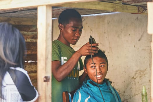 Ranohira, Madagascar - April 29, 2019: Unknown local boy getting his haircut from a friend at shop back room made from wooden boards. People in this part of Africa are poor, but cheerful.