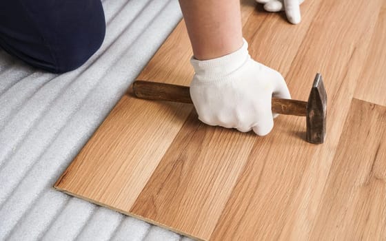 Installing laminated floor, detail on man hand in white gloves holding hammer over wooden tile, foam base layer under
