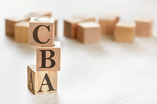 Three wooden cubes with letters CBA (means Cost Benefit Assessment), on white table, more in background, space for text in right down corner