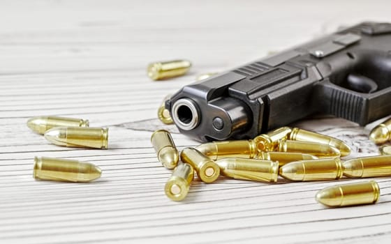 Closeup of modern dark gray gun with yellow brass bullets next to it on white wooden boards