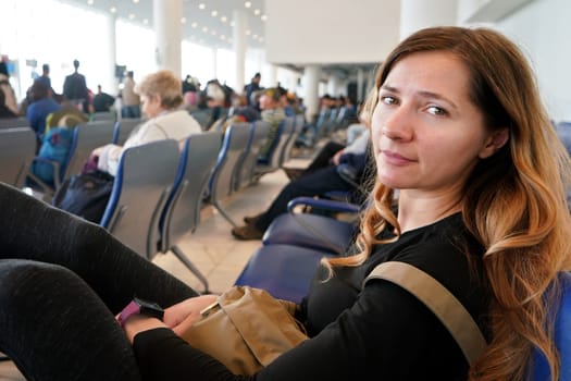 Young woman sitting at airport hall, looking tired after waiting couple of hours for connecting flight in early morning, more blurred passengers in background